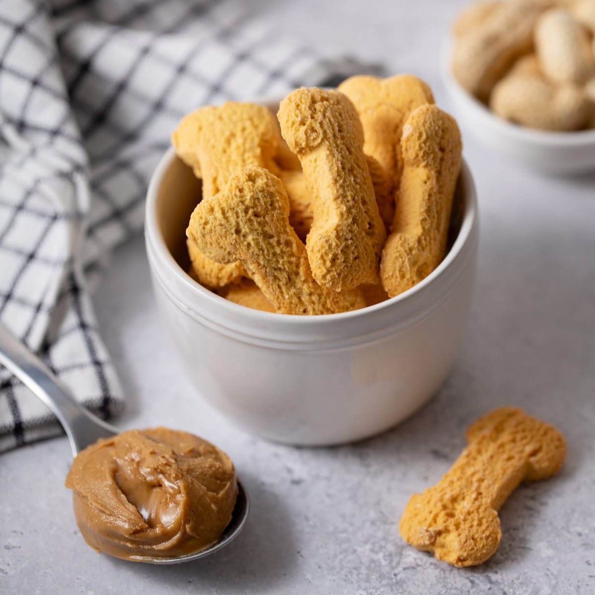 Dog treats shaped like bones in a white bowl on a gray surface with peanut butter and a checkered towel.
