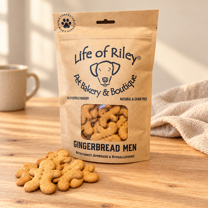 A stand-up kraft paper bag of Life of Riley Gingerbread Men dog treats sits on a wooden table, with several gingerbread-shaped treats spilled in front. The bag has a clear window showing the treats inside, and the background includes a softly blurred mug and fabric.